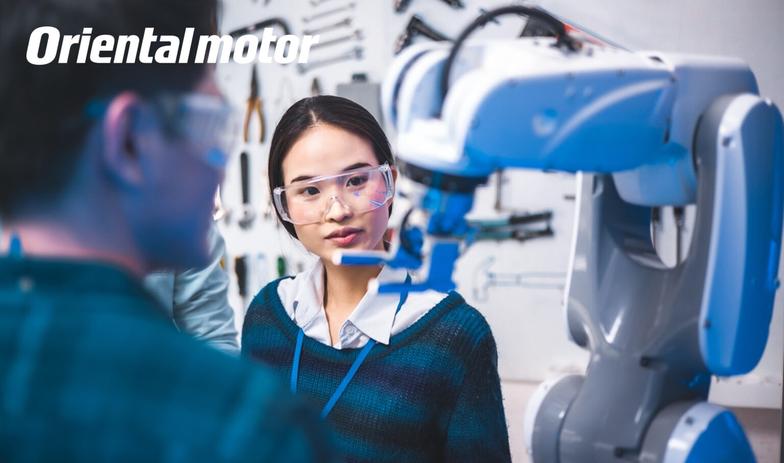 Frau mit Schutzbrille begutachtet Roboterarm bei der Arbeit, Indusrtieumgebung